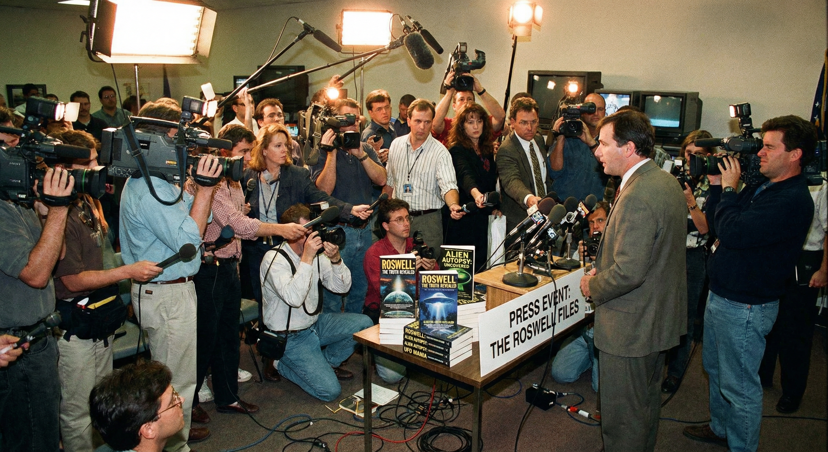 A 1990s media frenzy scene with television cameras, studio lights, and reporters crowded around a press event table displaying Roswell books